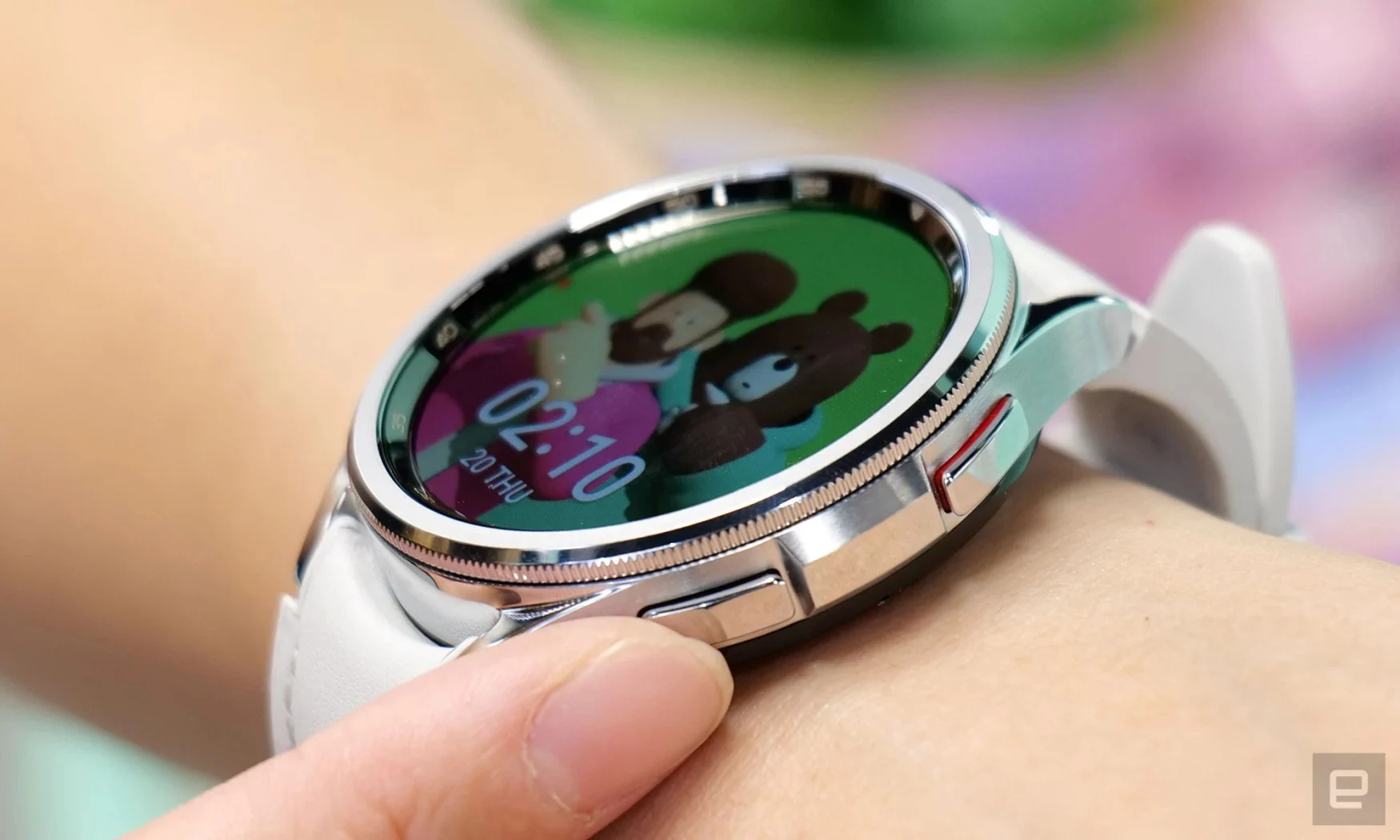 Una foto de primer plano de un reloj inteligente Samsung Galaxy Watch 6 en la muñeca de una persona.  Un dedo toca uno de los botones de hardware y aparece en la pantalla la imagen de un hombre con un osito de peluche.