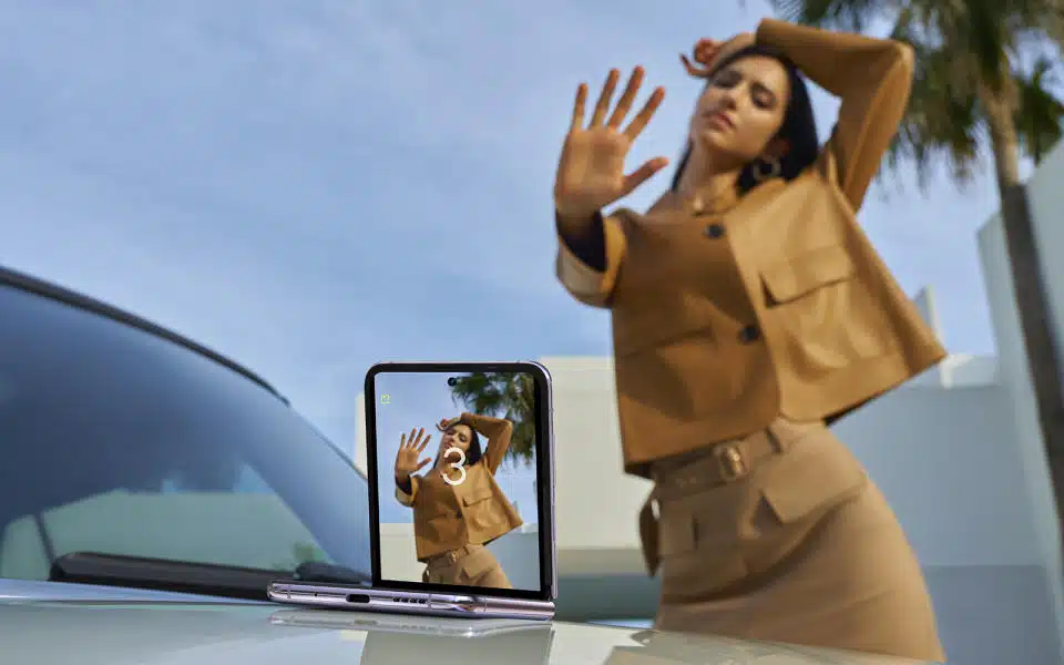 Un teléfono Tecno Phantom V Flip entreabierto colocado sobre el capó de un automóvil con la cámara principal mirando al modelo, quien posa con un gesto de la mano para disparar la foto de forma remota.