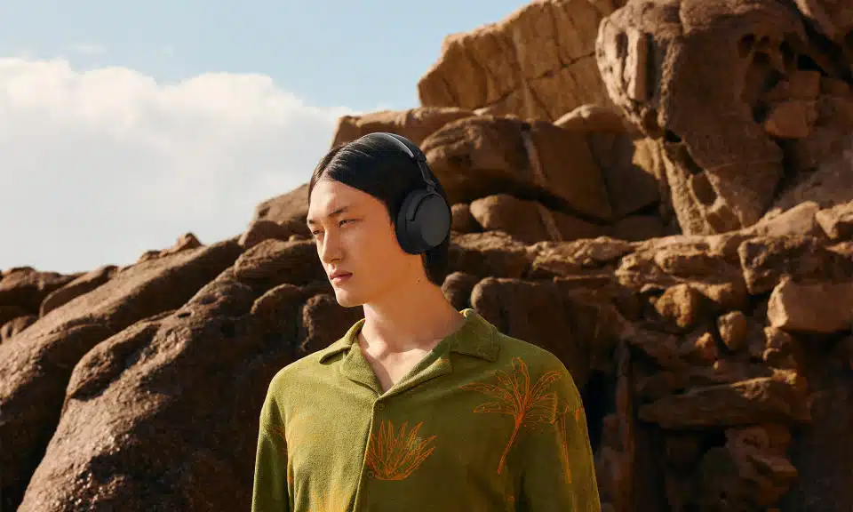 Fotografía comercial de los auriculares inalámbricos Sennheiser Accentum.  Un joven se encuentra en un terreno rocoso: detrás de él se levantan piedras.  Lleva una camisa verde con botones y motivos florales dorados/naranjas.